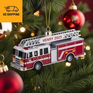May include: A red and white fire truck Christmas ornament with a ladder and the name "HENRY 2025" printed on it. The ornament is hanging from a gold string on a Christmas tree with red and gold ornaments.