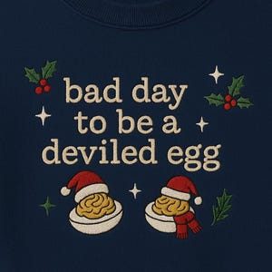 Op de afbeelding: Marineblauwe sweatshirt met geborduurde tekst "bad day to be a deviled egg". Het ontwerp bevat twee gevulde eieren met kerstmutsen en sjaals, omringd door hulst en sterren. Een feestelijk, humoristisch ontwerp.