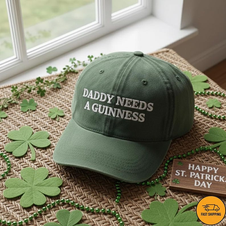 May include: A green baseball cap with the text "DADDY NEEDS A GUINNESS" in white. The cap is displayed on a woven surface with shamrock decorations and a "Happy St. Patrick's Day" sign.