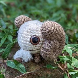 May include: A crocheted stuffed animal with brown ears and large brown eyes. The animal is white with a fluffy white beard. The animal is sitting on a rock with green leaves in the background.