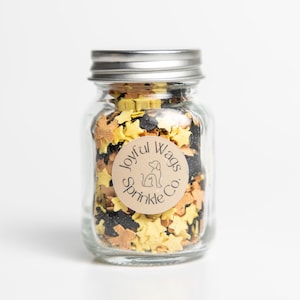 May include: A clear glass jar with a silver lid filled with star-shaped sprinkles in yellow, black, and tan colors. A round label on the jar reads "Joyful Wags Sprinkle Co." with a dog silhouette.