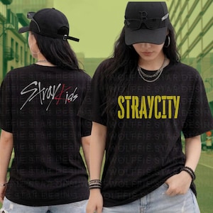 Puede incluir: Camisetas negras con el texto "STRAYCITY" en amarillo y "Stray Kids" en blanco y rojo. Las camisetas se llevan con gorras negras y collares plateados. El fondo es una calle de la ciudad.