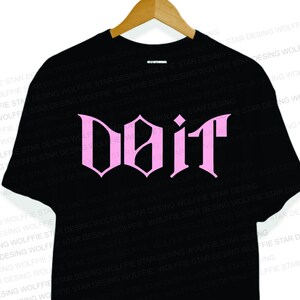 Puede incluir: Camiseta negra con la palabra "Doit" en rosa, con una fuente de estilo gótico. La camiseta está colgada en una percha de madera. El texto está centrado en la parte delantera.