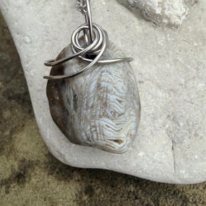 May include: A handcrafted pendant necklace featuring a polished, oval-shaped stone with a unique, patterned surface. The stone is wrapped with silver-colored wire and suspended from a delicate chain. The stone's colors range from light brown to off-white.