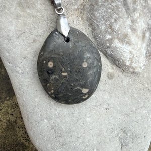 Könnte beinhalten: Dunkelgrauer Steinanhänger mit einer silberfarbenen Kette. Der Stein hat ein einzigartiges Muster aus helleren Flecken und Wirbeln. Der Anhänger hat eine Tropfenform und liegt auf einer hellgrauen Steinoberfläche. Ein natürliches, erdiges Accessoire.