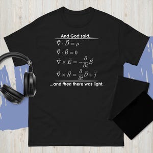 May include: Black t-shirt with white text. The text includes mathematical equations and the phrase "And God said... ...and then there was light.". A pair of black headphones and a black notebook are in the image.