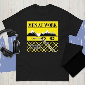 以下が含まれることがあります： 黒いTシャツ。黄色い長方形のデザインで、「MEN AT WORK」の文字と山脈のイラストが描かれています。山脈の下にはチェーンのデザインと「BUSINESS AS USUAL」の文字があります。
