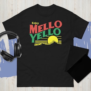 Puede incluir: Camiseta negra con las palabras "Enjoy Mello Yello" en rojo y verde, con un gráfico de sol amarillo sobre una cordillera. La camiseta tiene mangas cortas y cuello redondo.
