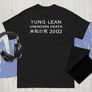 May include: Black t-shirt with white text reading "YUNG LEAN UNKNOWN DEATH 未知の死 2002". The image also shows black headphones and a black notebook. The t-shirt is made of a soft material.