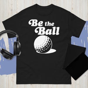 May include: Black t-shirt with the text "Be the Ball" in white script above a white golf ball graphic. The golf ball has detailed dimples and a shadow. Headphones and a black notepad are visible.