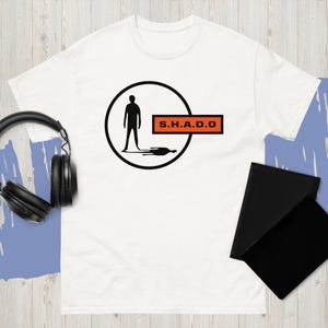 May include: White t-shirt with a black silhouette of a person inside a black circle, with a shadow. The text "S.H.A.D.O" is in an orange rectangle. Black headphones and two black notebooks are visible.