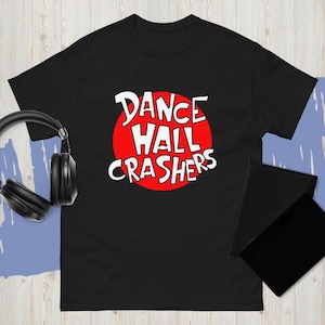 Może przedstawiać: Czarny t-shirt z białym napisem "DANCE HALL CRASHERS" na czerwonym okręgu. Koszulka ma krótkie rękawy. Czarne słuchawki leżą po lewej stronie koszulki.