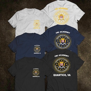 Puede incluir: Una colección de camisetas de manga corta en blanco, azul marino y negro. Cada camiseta presenta el texto "FBI ACADEMY" y un emblema circular. Algunas camisetas también muestran "QUANTICO, VA" y el sello del Departamento de Justicia.