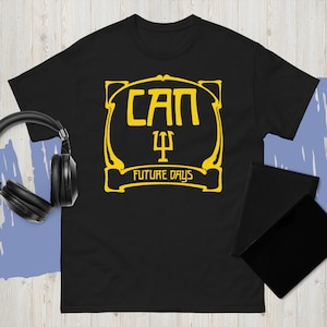 May include: Black t-shirt with a yellow graphic. The graphic features the word "CAN" in stylized letters, a symbol, and the words "FUTURE DAYS". A pair of black headphones and a black notebook are visible.