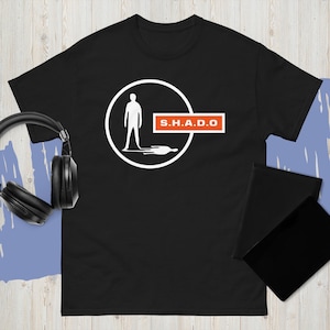 May include: Black t-shirt with a white graphic of a person inside a circle, with a shadow below. The text "S.H.A.D.O" is in a red rectangle. Headphones and a black notebook are visible.