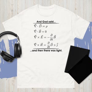 May include: White t-shirt with black text that reads "And God said..." followed by mathematical equations and the phrase "...and then there was light." A pair of black headphones and two black notebooks are also visible.