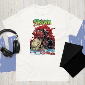 Könnte beinhalten: Weißes T-Shirt mit einem Comic-Grafik von Spawn, einer Superheldenfigur. Die Grafik enthält das Wort "Spawn" in Grün und Rot sowie eine detaillierte Illustration der Figur in einem roten Umhang. Das Shirt ist aus weichem Material.