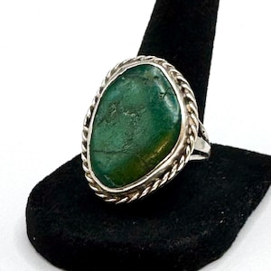 May include: A silver ring featuring a large, irregular, oval-shaped green stone with natural dark veining. The stone is set within a twisted rope-style silver border, mounted on a simple, polished silver band that tapers toward the back.