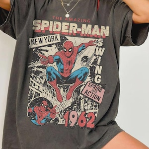 May include: A charcoal gray short-sleeve t-shirt featuring a graphic of Spider-Man in a dynamic pose. The print includes the text THE AMAZING SPIDER-MAN, NEW YORK, SPRING INTO ACTION, and the year 1962. The design has a distressed, vintage-inspired look.
