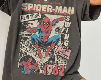 Può includere: Una t-shirt a maniche corte color grigio antracite con una grafica di Spider-Man in una posa dinamica. La stampa include il testo THE AMAZING SPIDER-MAN, NEW YORK, SPRING INTO ACTION e l'anno 1962. Il design ha un aspetto vissuto e vintage.