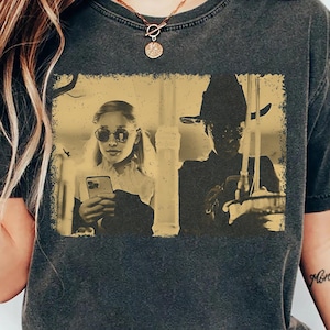 Può includere: Una t-shirt a maniche corte color grigio antracite con una stampa grafica rettangolare in tonalità seppia. La grafica mostra due figure: una persona con occhiali da sole che tiene uno smartphone e un'altra con un cappello a tesa larga.