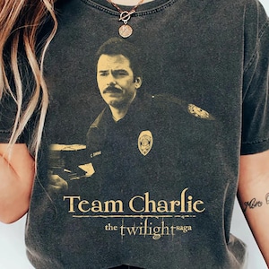 May include: Dark gray t-shirt featuring a sepia-toned image of a man in a police uniform with a mustache. The text "Team Charlie" and "the twilight saga" are printed in gold lettering below the image.