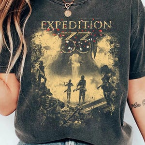 Può includere: Una t-shirt a maniche corte grigio antracite con una stampa grafica che riporta il testo "EXPEDITION 33" in lettere dorate. Il design raffigura un gruppo di figure in un paesaggio boschivo con una cascata, arricchito da petali rossi.