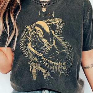 May include: A charcoal grey short-sleeve t-shirt featuring a yellow graphic print of the Alien creature from the film franchise. The design includes the word ALIEN in a stylised font above the creature, which is depicted with its tail coiled.