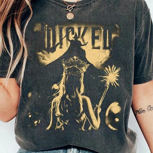 Könnte beinhalten: Dunkelgraues T-Shirt mit dem Wort "WICKED" in Gold, über einer Silhouette einer Hexe und einer gekrönten Frau mit einem Zauberstab. Das Design hat einen Vintage-Look.