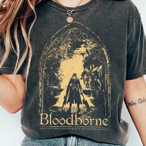 Peut inclure: T-shirt gris foncé avec un graphisme jaune et noir du jeu vidéo Bloodborne. Le graphisme représente une silhouette en manteau long dans une arche gothique. Le texte "Bloodborne" et "May you find your worth in the waking world" sont en dessous.