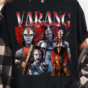Pode incluir: Uma t-shirt preta de manga curta com uma impressão gráfica com o texto VARANG numa fonte com gradiente vermelho e branco. O design apresenta cinco figuras humanoides de pele azul com marcas faciais e toucados vermelhos num layout de colagem.