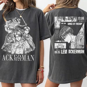 May include: Dark gray t-shirt featuring a black and white graphic of anime characters with the text "ACKERMAN" on the front. The back of the shirt has a graphic with the text "WINGS OF FREEDOM" and "LEVI ACKERMAN".