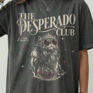 May include: A charcoal grey t-shirt featuring a graphic print of a cat wearing a crown and sunglasses, sitting behind a bowl of cherries. The text reads THE DESPERADO CLUB and SO FUN IT HURTS, accompanied by a dagger illustration and small star accents.