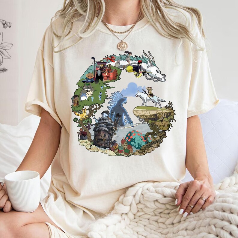 May include: Cream-colored t-shirt featuring a colorful graphic design of a dragon and various animated characters. The design is in the shape of a letter "G". The shirt is made of a soft material.