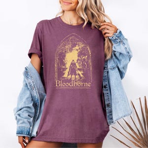 May include: A maroon short-sleeved t-shirt featuring a yellow graphic print of a gothic archway framing a silhouetted figure. The text reads Bloodborne and May you find your worth in the waking world. The shirt is worn under a light blue denim jacket.