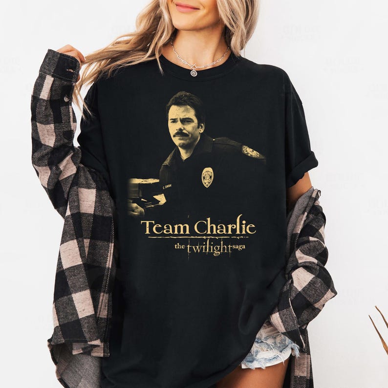 May include: Black t-shirt featuring a sepia-toned image of a man in a police uniform. The text "Team Charlie" is printed above "the twilight saga" in a gold color. The shirt is worn by a person.