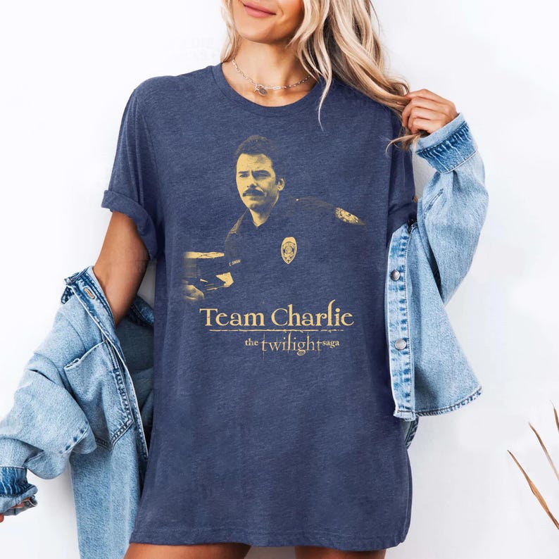 May include: Navy blue t-shirt featuring a gold image of a man with the text "Team Charlie the twilight saga". The shirt is a crew neck and has short sleeves. The shirt is worn by a person wearing a denim jacket.