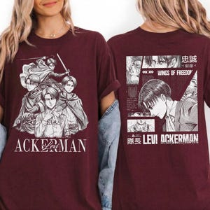 May include: Maroon t-shirt featuring a manga-style illustration of characters with swords and capes. The front of the shirt displays the word "ACKERMAN" in white. The back of the shirt has a manga panel with the text "WINGS OF FREEDOM" and "LEVI ACKERMAN".