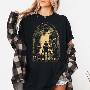 May include: A black t-shirt featuring a gold graphic of a character standing in a gothic archway. The text reads Bloodborne and May You Find Your Worth In The Waking World. The shirt is worn under a black and gray plaid long-sleeve button-down shirt.