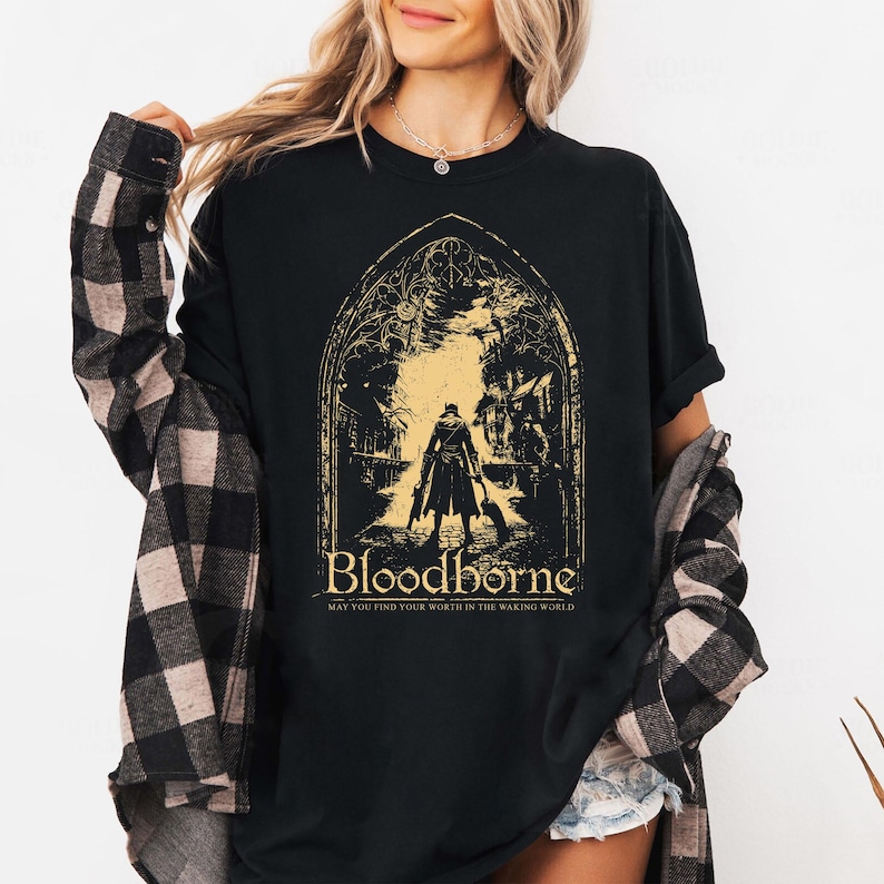 May include: A black t-shirt featuring a gold graphic of a character standing in a gothic archway. The text reads Bloodborne and May You Find Your Worth In The Waking World. The shirt is worn under a black and gray plaid long-sleeve button-down shirt.