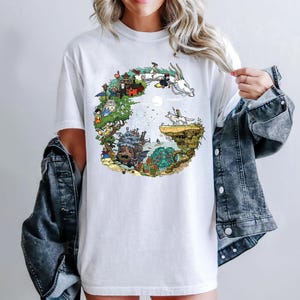 May include: White t-shirt with a vibrant graphic depicting a fantasy scene with various characters and creatures. The design includes a dragon, a castle, and other imaginative elements. The t-shirt is made of a soft fabric.