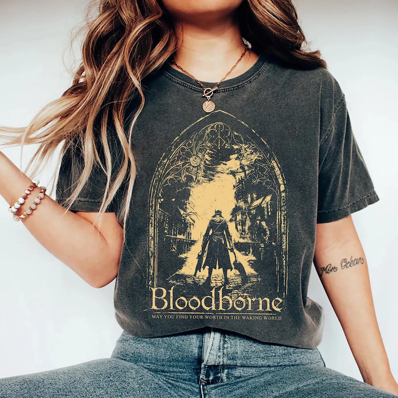 May include: A charcoal gray short-sleeve t-shirt featuring a yellow graphic print of a figure in a long coat standing within a gothic archway. The text "Bloodborne" and "May you find your worth in the waking world" is printed below the illustration.