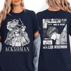 May include: Navy blue t-shirt featuring a black and white graphic of anime characters. The front of the shirt displays a group of characters with the text "ACKERMAN". The back of the shirt has a manga-style design with the text "WINGS OF FREEDOM" and "LEVI ACKERMAN".