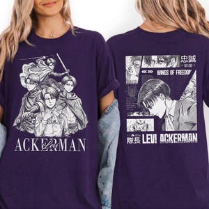 May include: A dark purple t-shirt featuring a manga-style illustration of several characters. The front of the shirt has the word "ACKERMAN" in white. The back of the shirt has a manga panel with the words "WINGS OF FREEDOM" and "LEVI ACKERMAN".