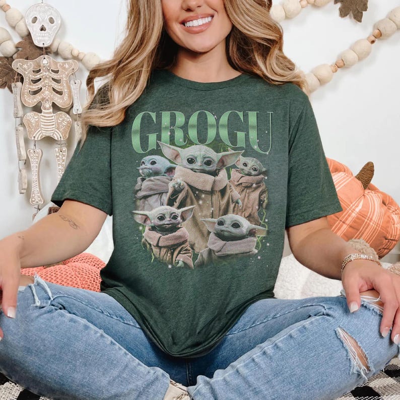 Puede incluir: Camiseta verde oscuro con la palabra "GROGU" en texto verde sobre una ilustraci&oacute;n de Grogu de The Mandalorian. El gr&aacute;fico incluye m&uacute;ltiples im&aacute;genes de Grogu en varias poses. La camiseta es de estilo manga corta.