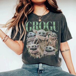 Puede incluir: Camiseta gris oscuro con la palabra "GROGU" en verde sobre un gr&aacute;fico de Grogu, tambi&eacute;n conocido como Baby Yoda, de The Mandalorian. El gr&aacute;fico muestra a Grogu en varias poses. La camiseta es de estilo vintage.