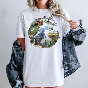 May include: White t-shirt featuring a colorful graphic design of a fantasy landscape with various characters and elements. The design is arranged in a circular shape. The shirt is made of a soft material.