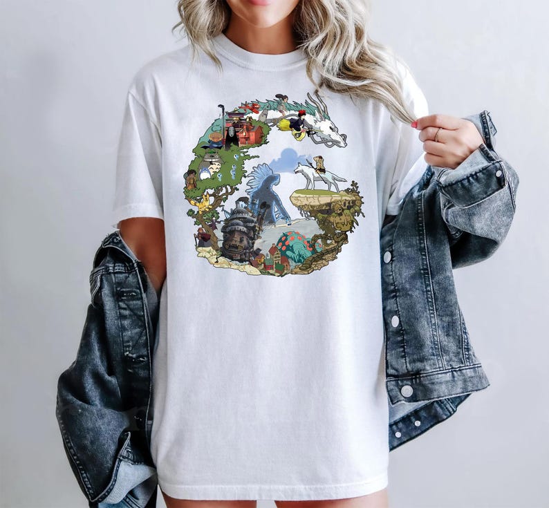 May include: White t-shirt featuring a colorful graphic design of a fantasy landscape with various characters and elements. The design is arranged in a circular shape. The shirt is made of a soft material.