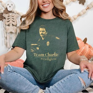 May include: Dark green t-shirt featuring a sepia-toned image of a man in a police uniform. The text "Team Charlie" and "the twilight saga" are printed in gold below the image. The shirt is a short-sleeved crew neck.