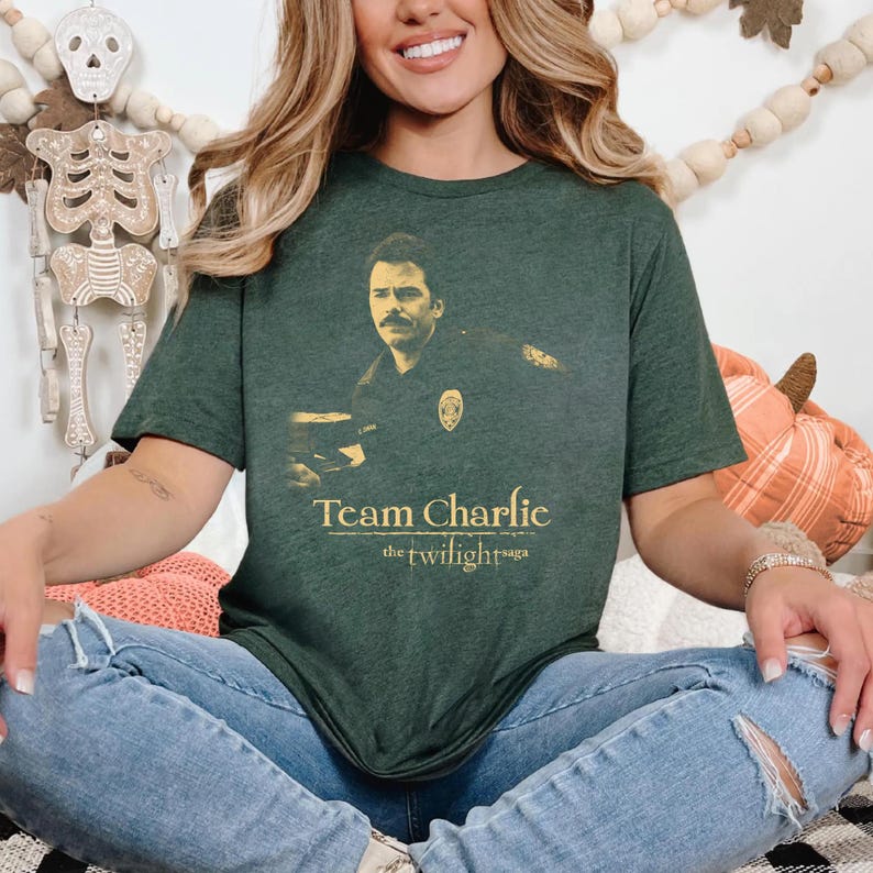 May include: Dark green t-shirt featuring a sepia-toned image of a man in a police uniform. The text "Team Charlie" and "the twilight saga" are printed in gold below the image. The shirt is a short-sleeved crew neck.